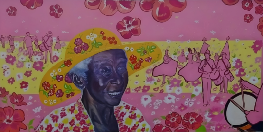 Um suave sorriso, chapéu colorido e vestes floridas, a figura principal com olhar feliz e contemplativo é Francisca Ramos dos Santos, matriarca do Marabaixo conhecida como “Tia Chiquinha”. Na cena, a dama da cultura popular amapaense participa da retirada do mastro, cortejo festivo que celebra a alegria, tradição e o legado de resistência em suas bandeiras, caixas, tocadores e dançadeiras.